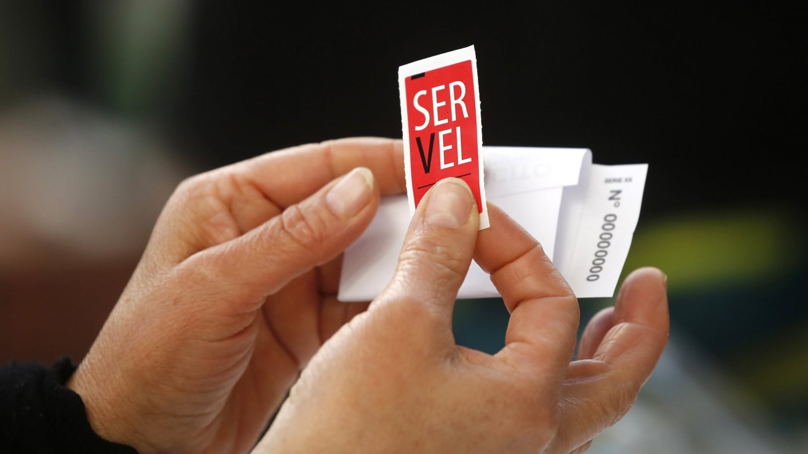 Manos sujetando un sticker del Servel y un voto doblado.-