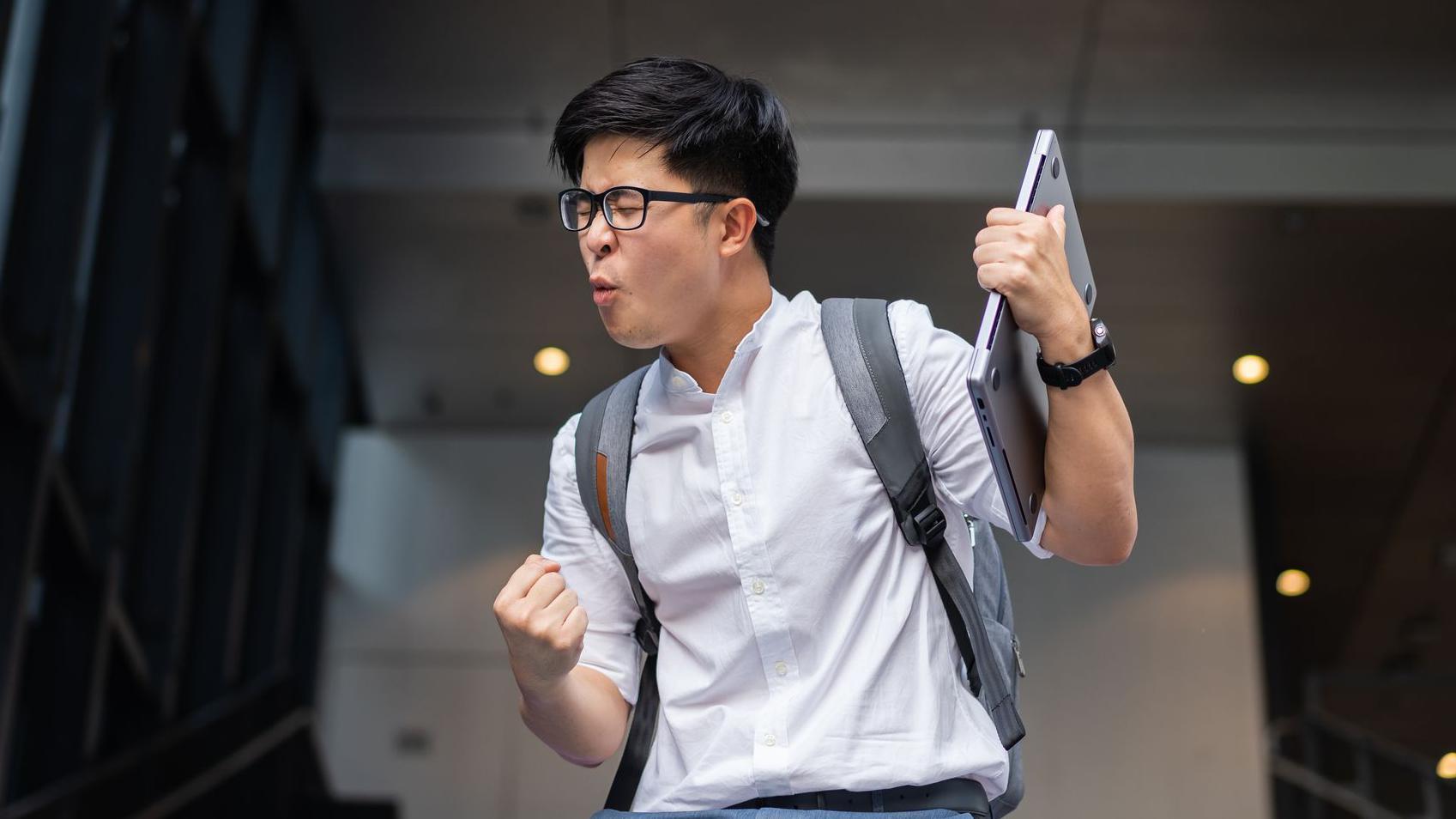 Un hombre con mochila a sus espaldas y una computadora en una mano hace un gesto de satisfacción y alegría con la cara y el otro brazo.